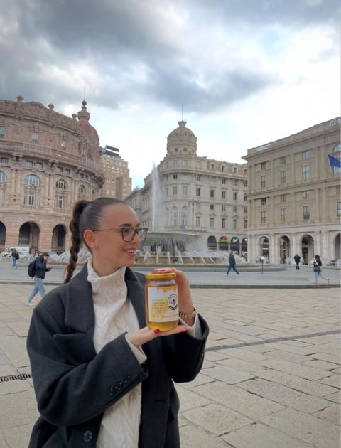 Woman wearing glasses and black blazer holding a honey jar in a European city square with classical buildings